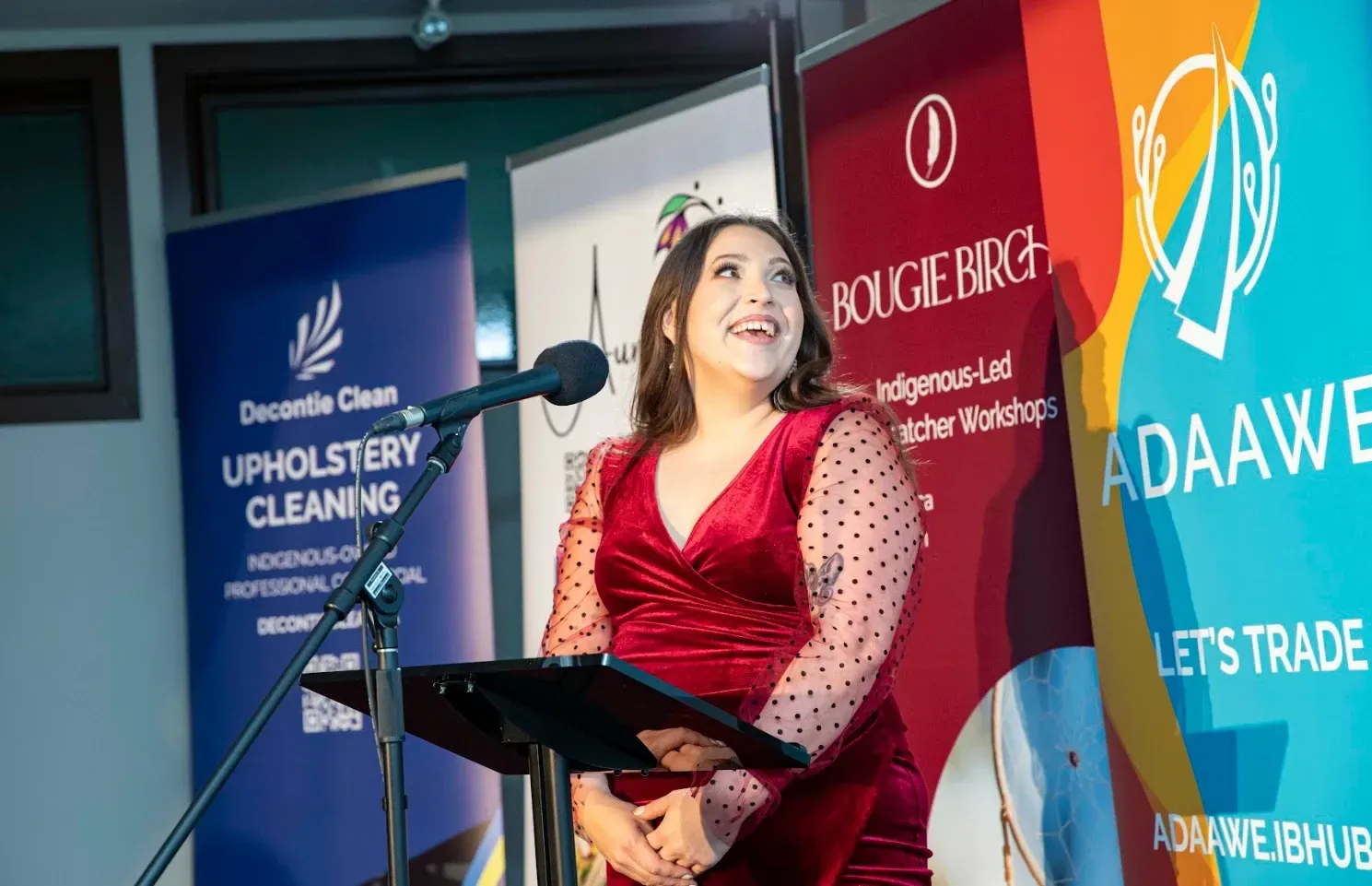 Woman in red dress speaking at a podium, smiling. Event with banners featuring logos.