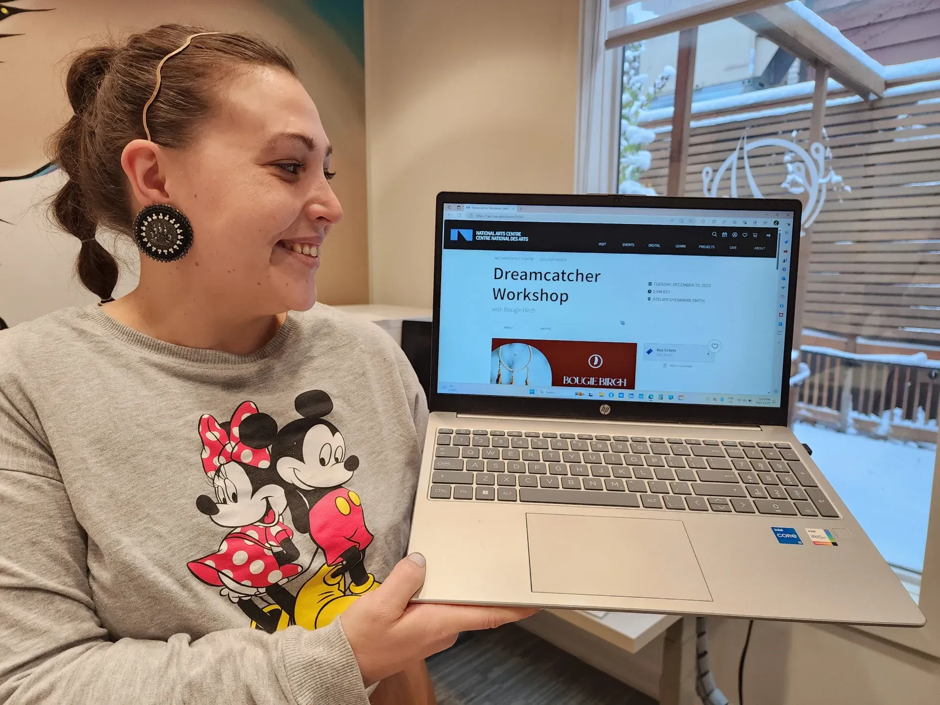 Woman holding a laptop, smiling. Laptop displays a website, possibly a school. Snowy window in the background.