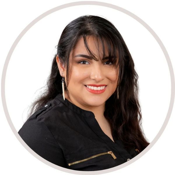 Woman with dark hair, bangs, smiling, wearing a black shirt with gold zippers and dangling earrings.