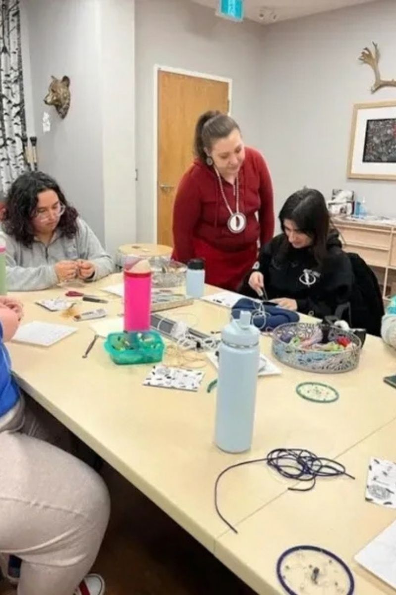 Group crafting with an instructor, seated at a long table. Beads, string, and supplies are scattered about.