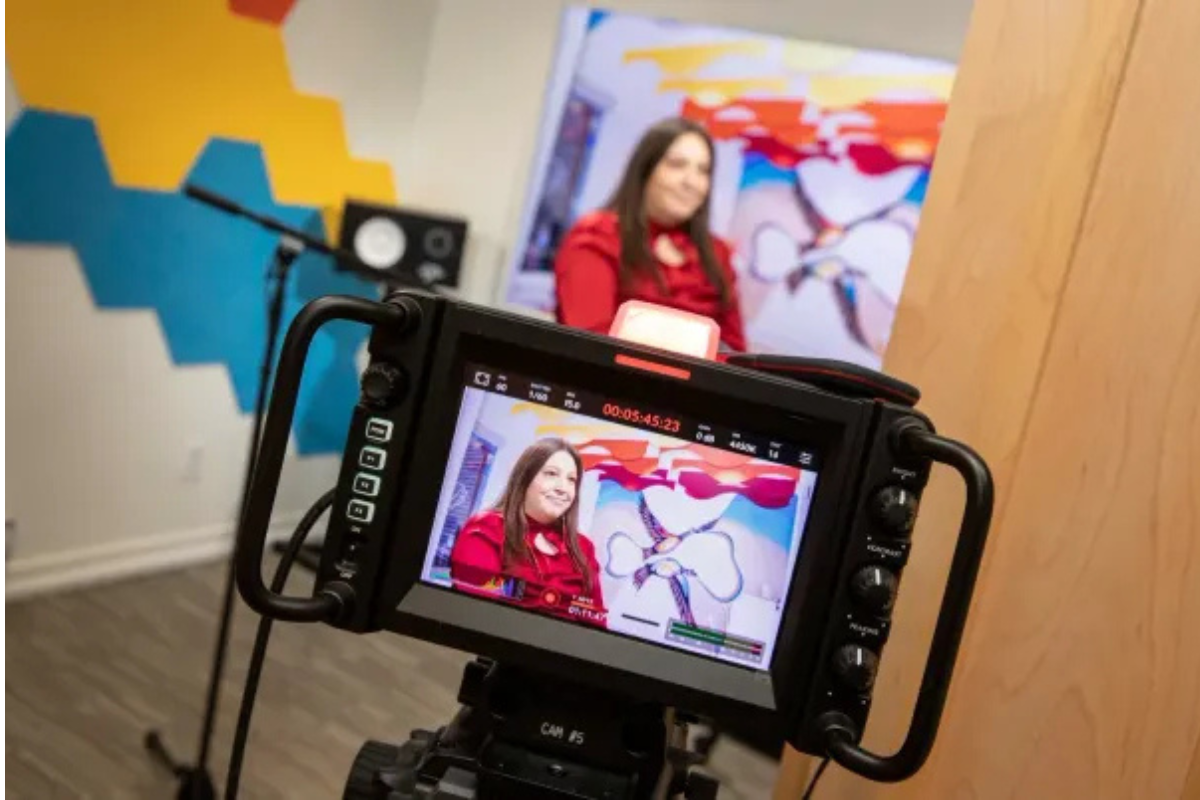 A person being filmed in a studio with colorful wall art and camera equipment. The person is wearing red.