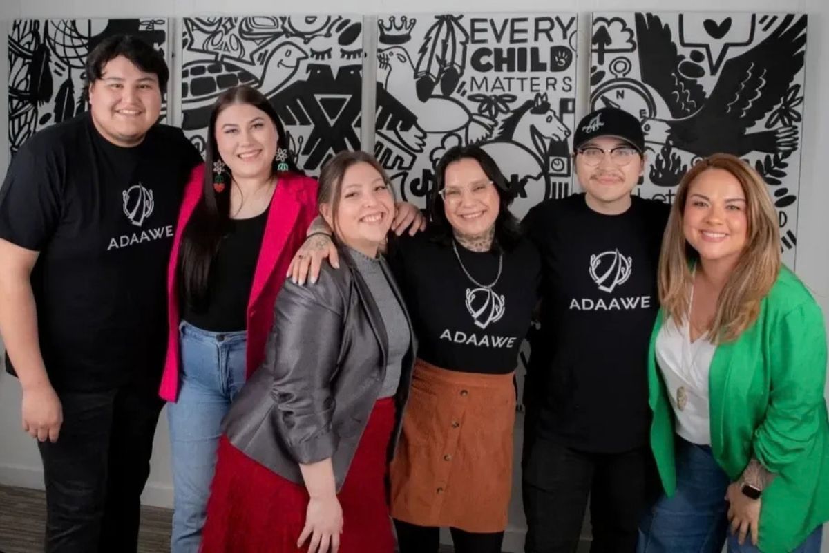 Group of six people smiling, posing in front of a black and white mural. They wear black ADAWE shirts or hats.