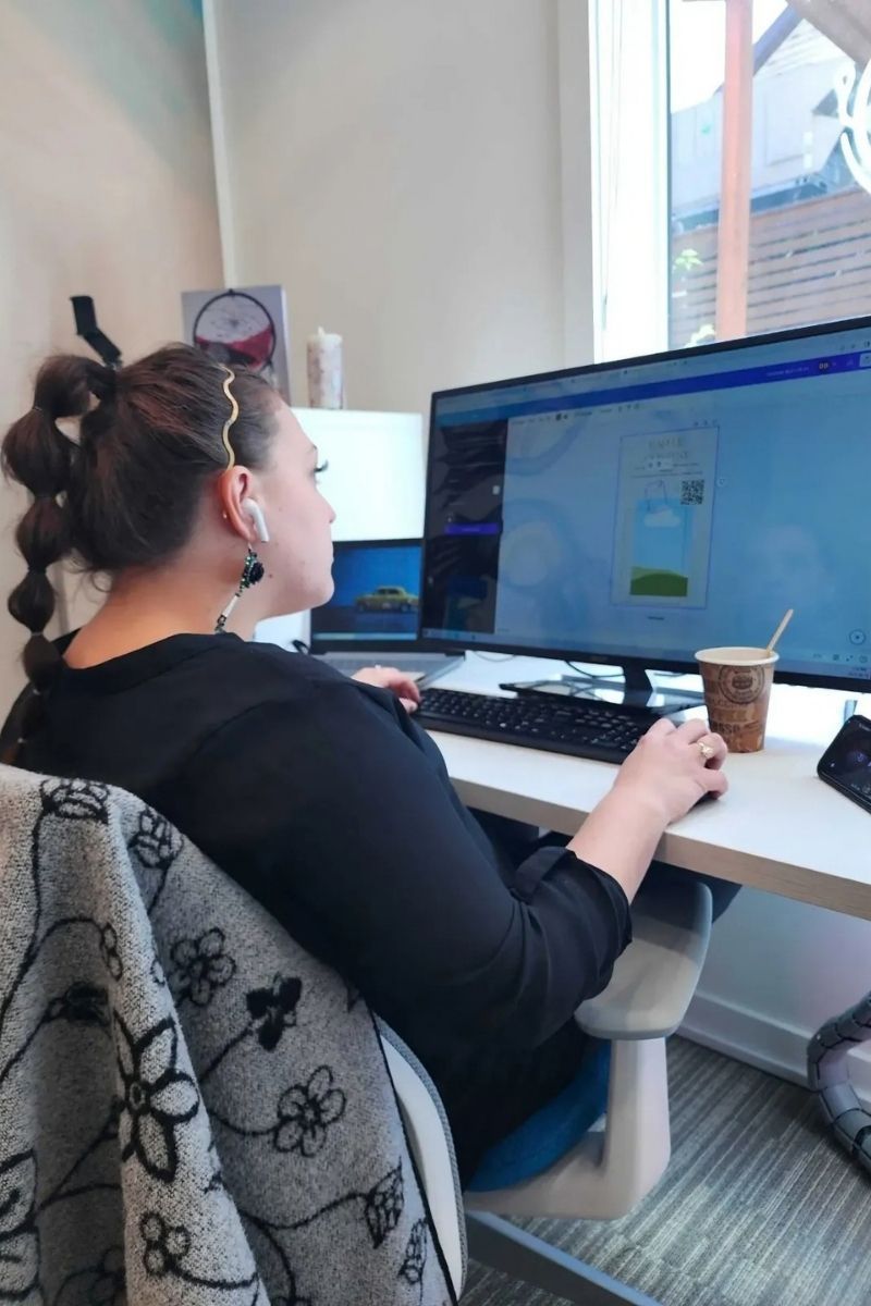 Woman at computer, right hand on mouse.  Wearing headphones, seated at a desk near a window. Coffee cup and phone present.