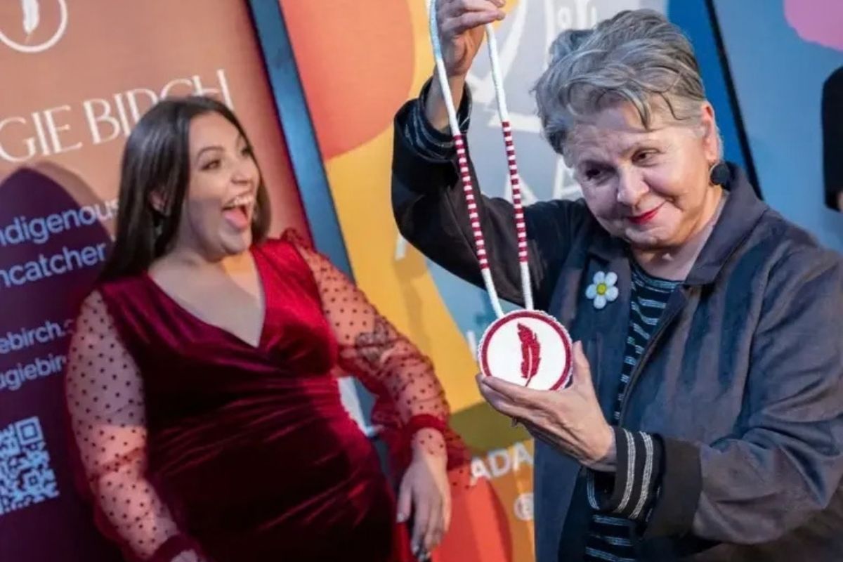 Woman holds medallion with feather design; another woman reacts excitedly.