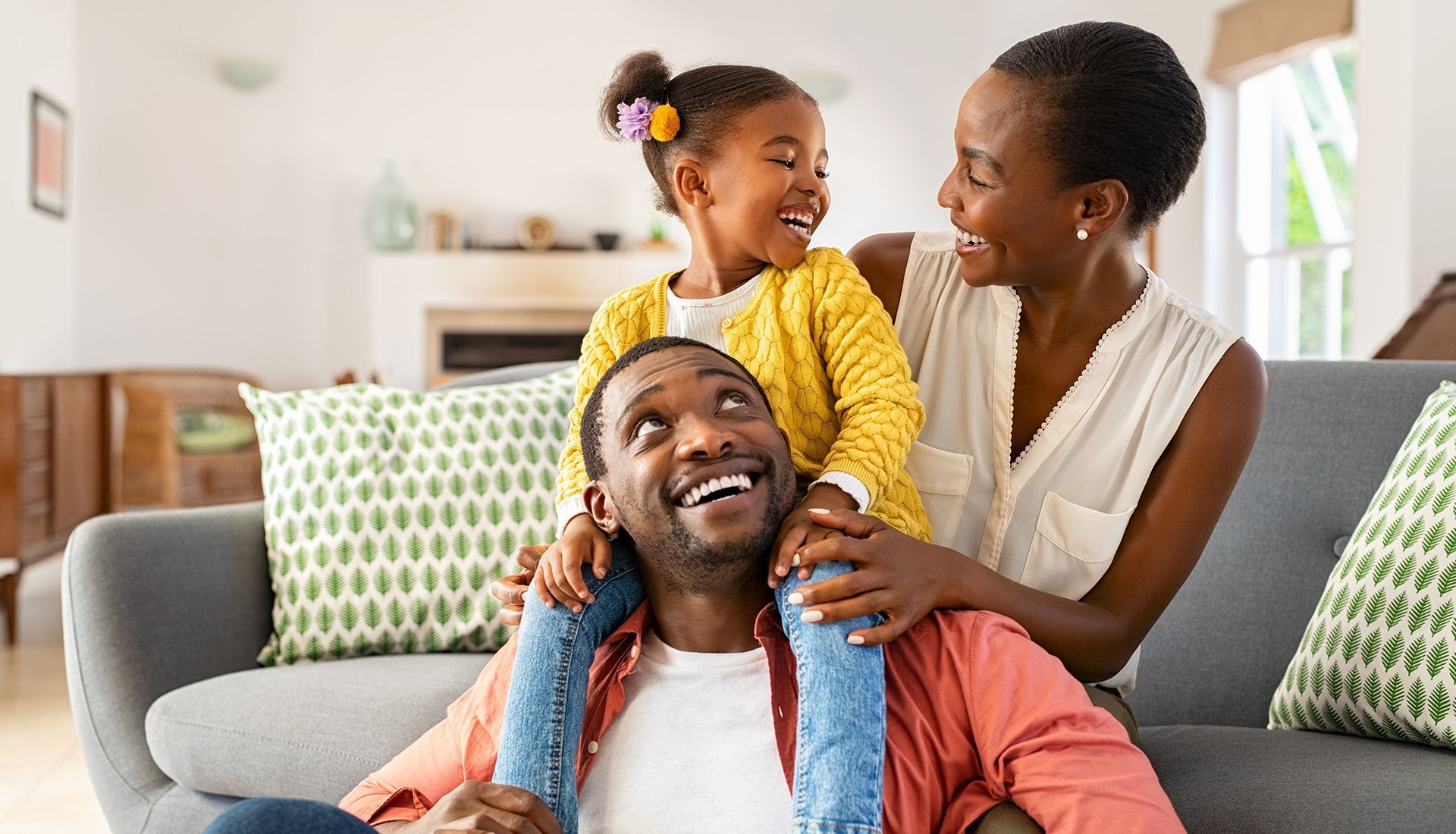 Smiling Black family: father with daughter on shoulders, mother beside them on couch.
