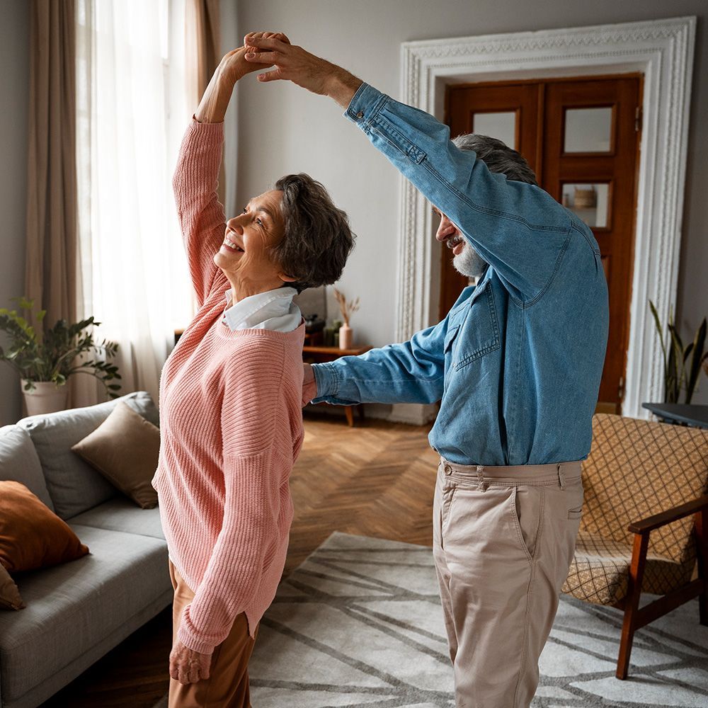 Elderly couple dancing in a living room. The woman is smiling and the man is supporting her.