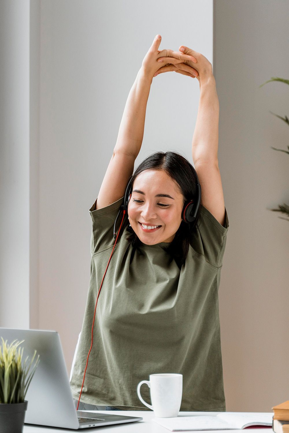Woman in green shirt stretches arms up while seated at a desk with a laptop and mug.