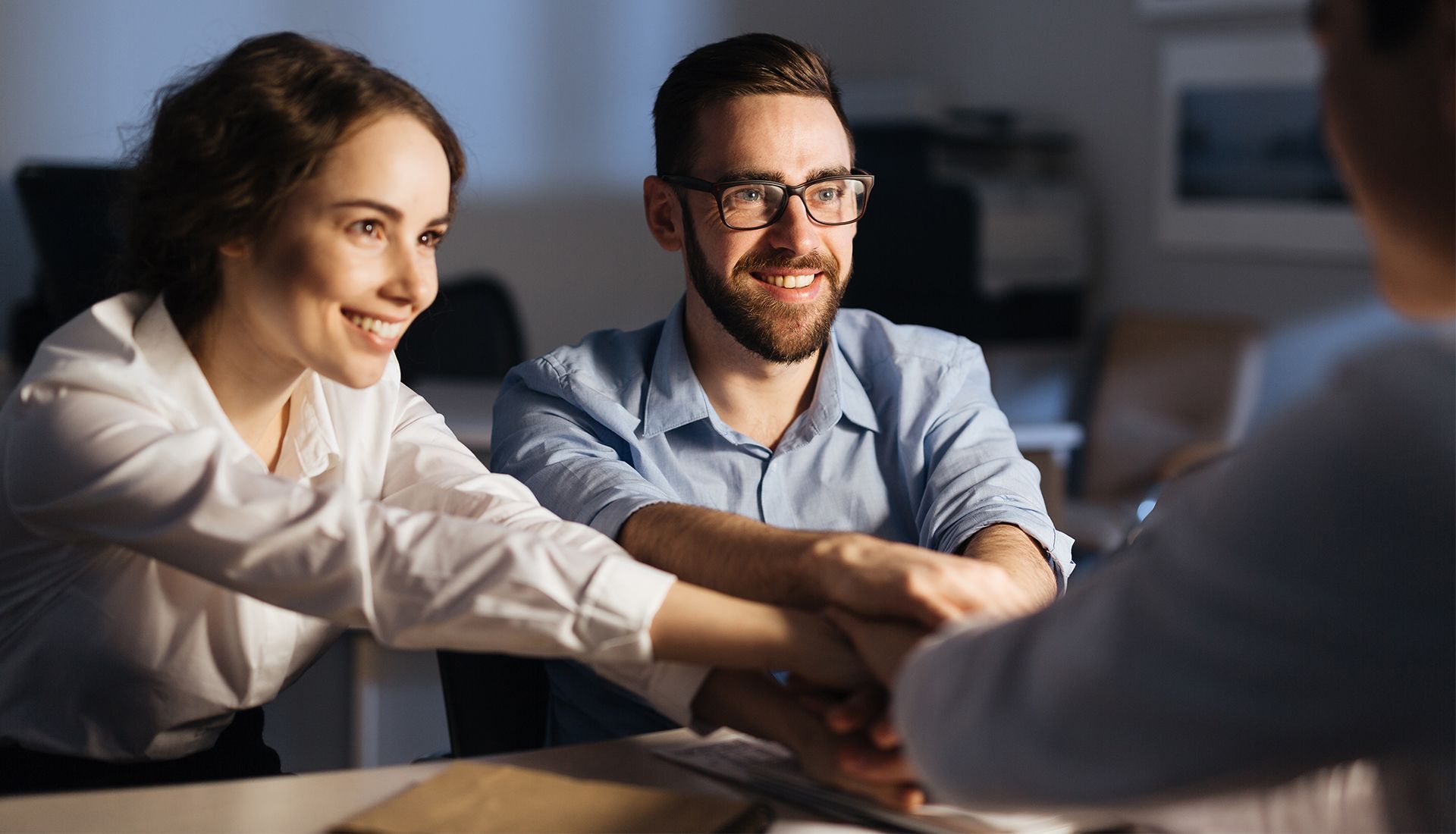 Couple with hands stacked on top of another person's; smiling, office setting.