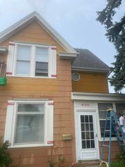 Two-story house exterior with tan siding, white trim, and a worker on a blue ladder by the front door.