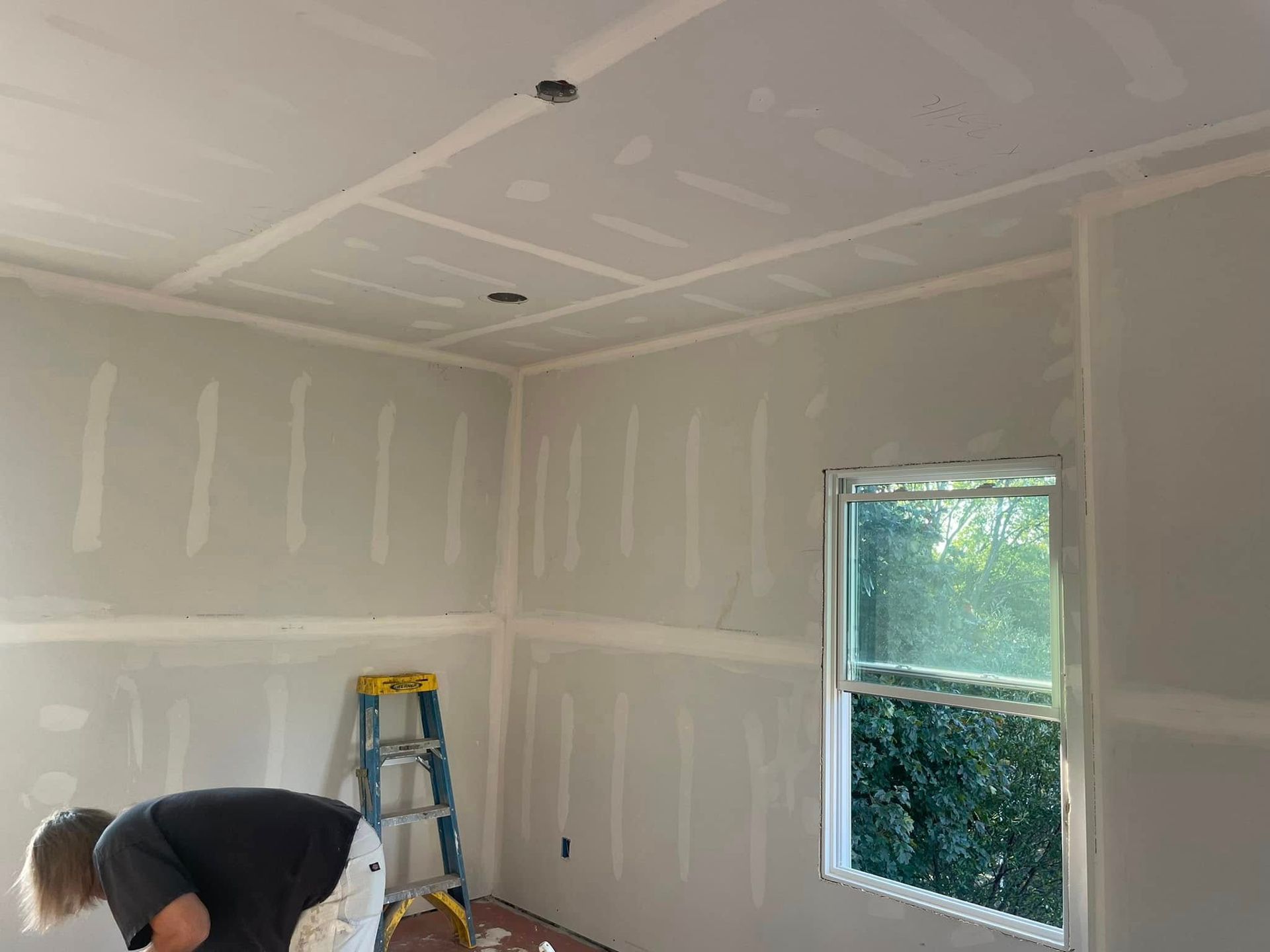 Unfinished room with drywall seams, a ladder, and a person working near the floor by a window.