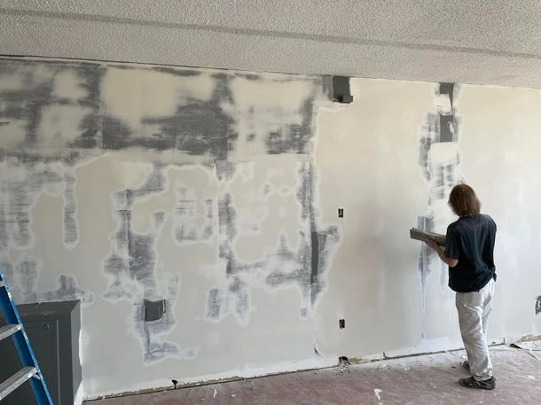 Person painting a white wall in a room under renovation, with patches of drywall repair and a ladder nearby