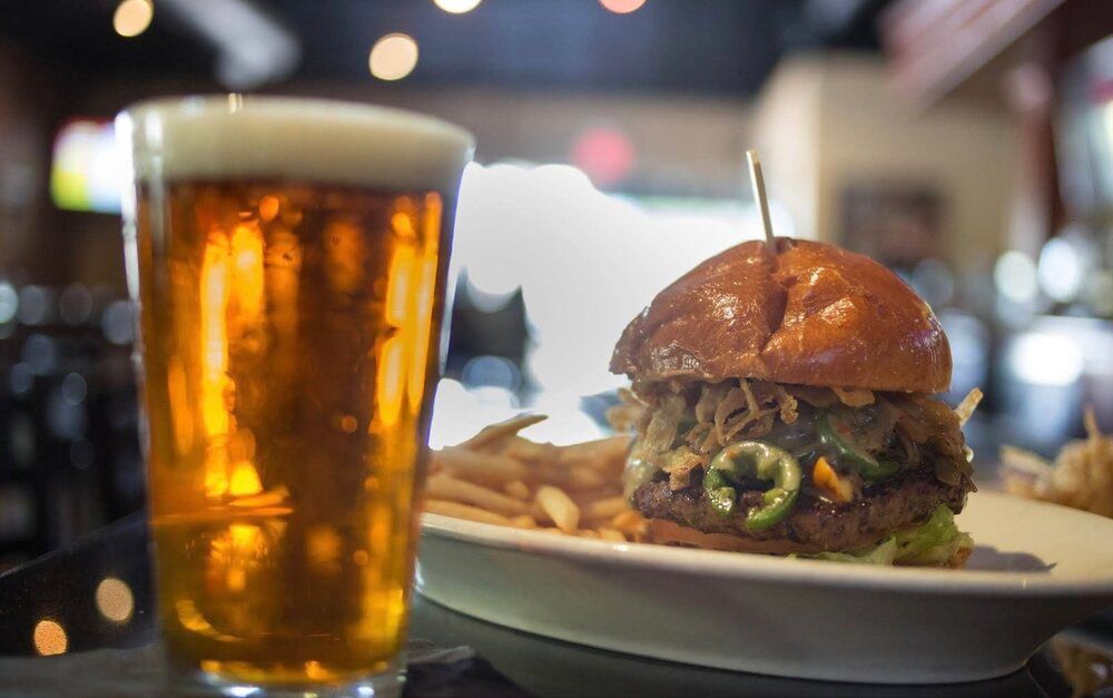 A beer glass beside a burger and fries on a restaurant table, with warm blurred lights in the background.