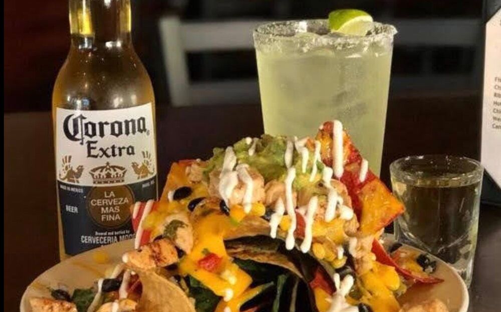Plate of nachos with drizzled sauces beside a Corona Extra beer and a lime drink on a table