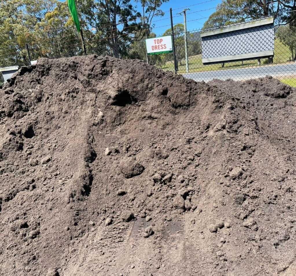 Pile of dark brown soil outdoors with a sign in the background that reads 
