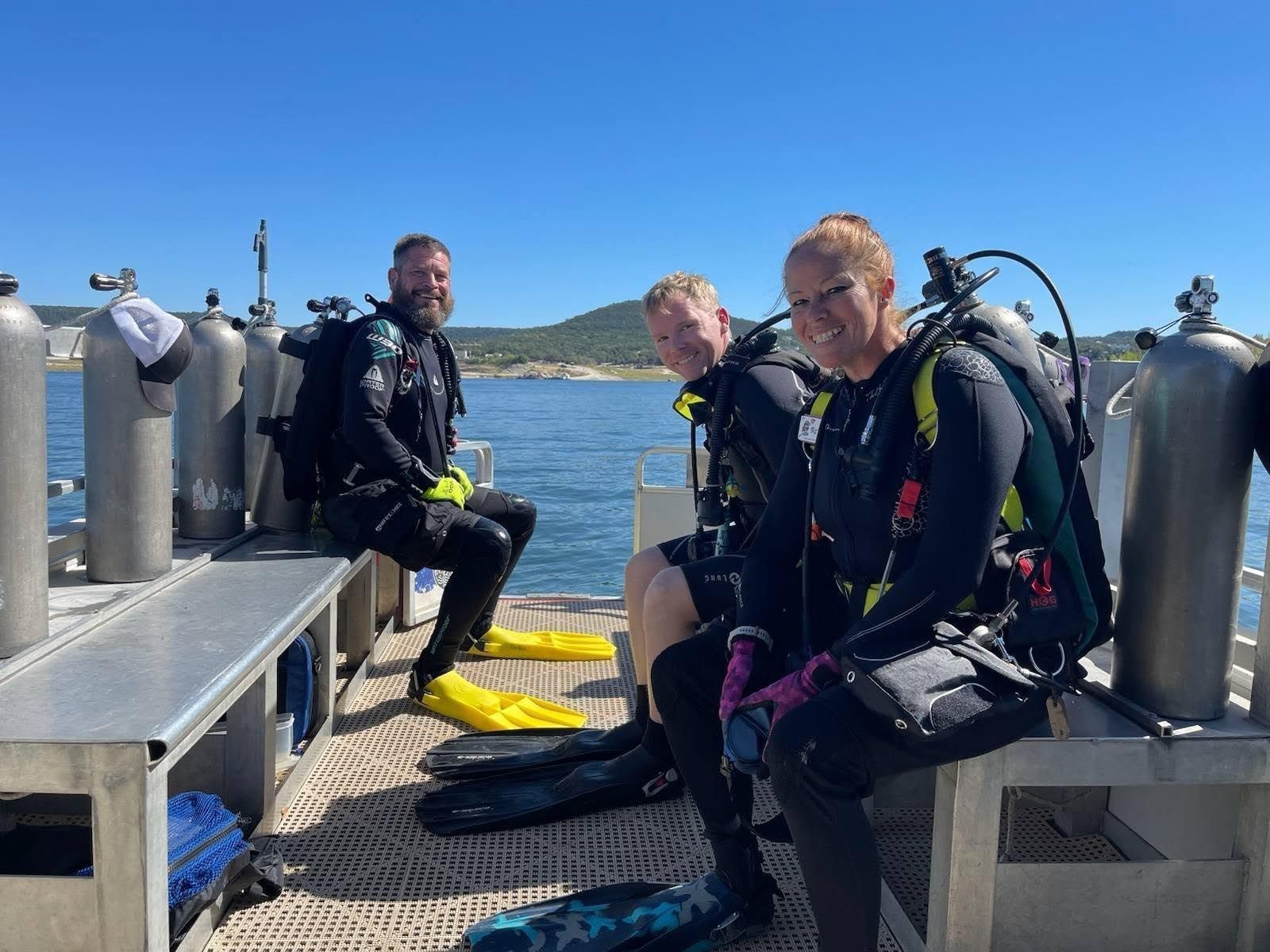 Three scuba divers seated on a boat, preparing to dive. Blue sky, ocean visible.