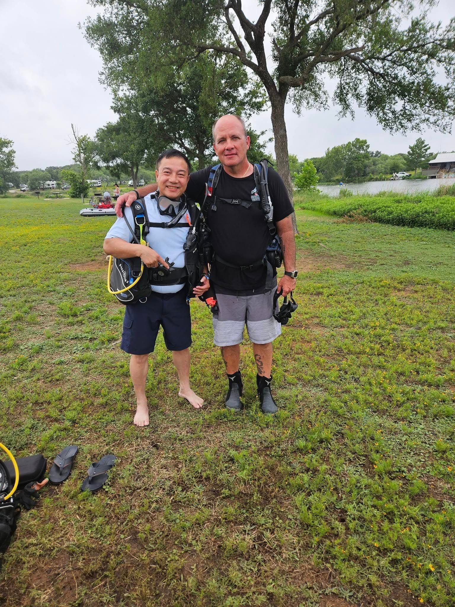Two men in scuba gear stand on grass by water. One man has his arm around the other's shoulder; both smiling.