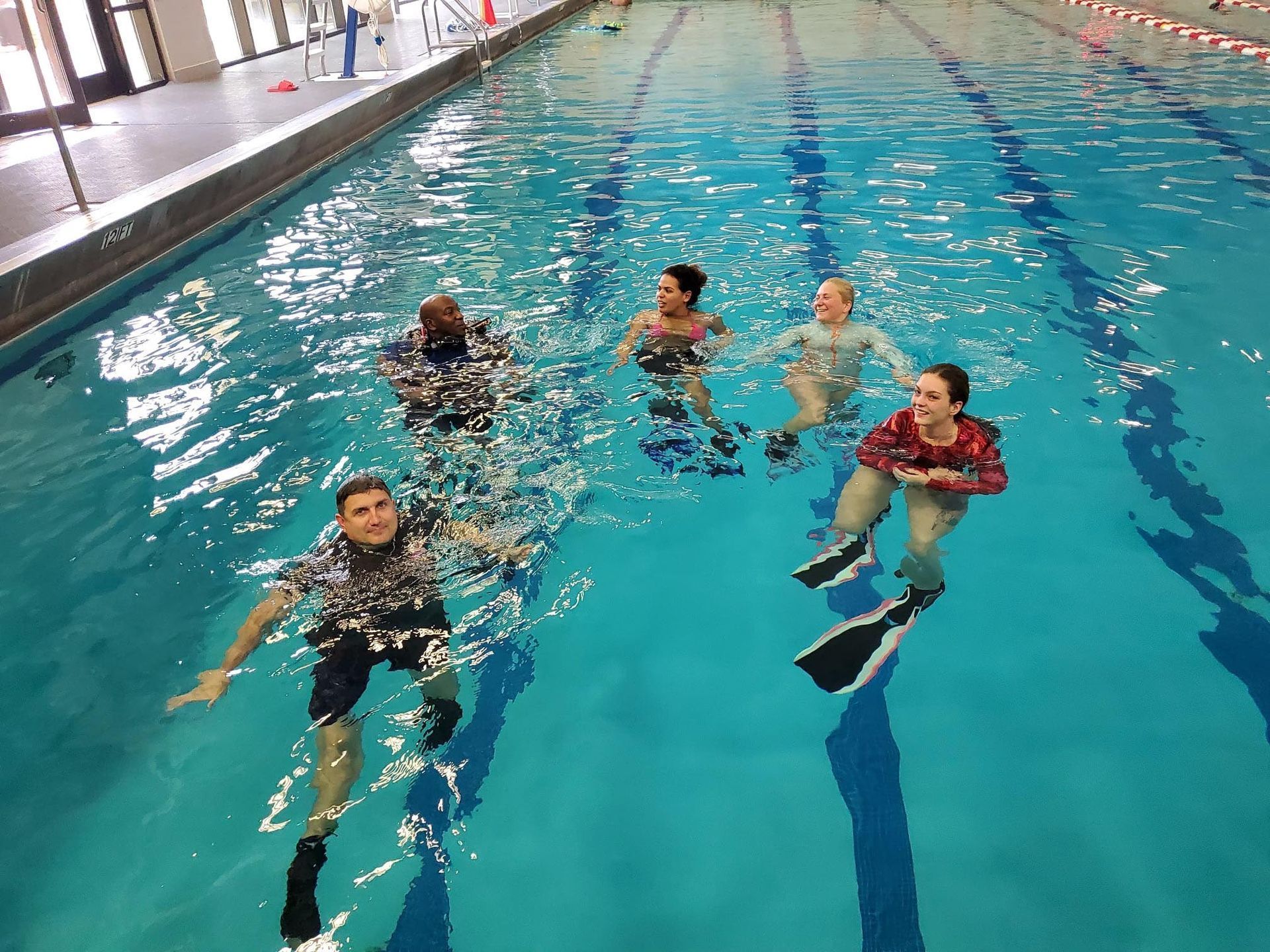 People in a pool wearing fins, exercising. Bright blue water. Indoors, with windows and red and white barriers.