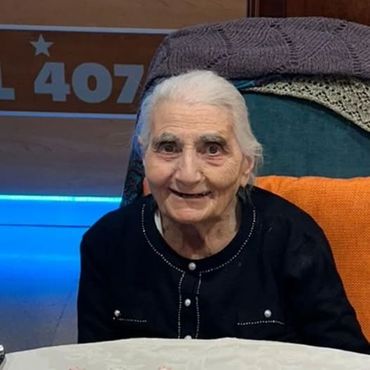 Una donna anziana sorride; è seduta in casa davanti a una luce blu e a un cartello.