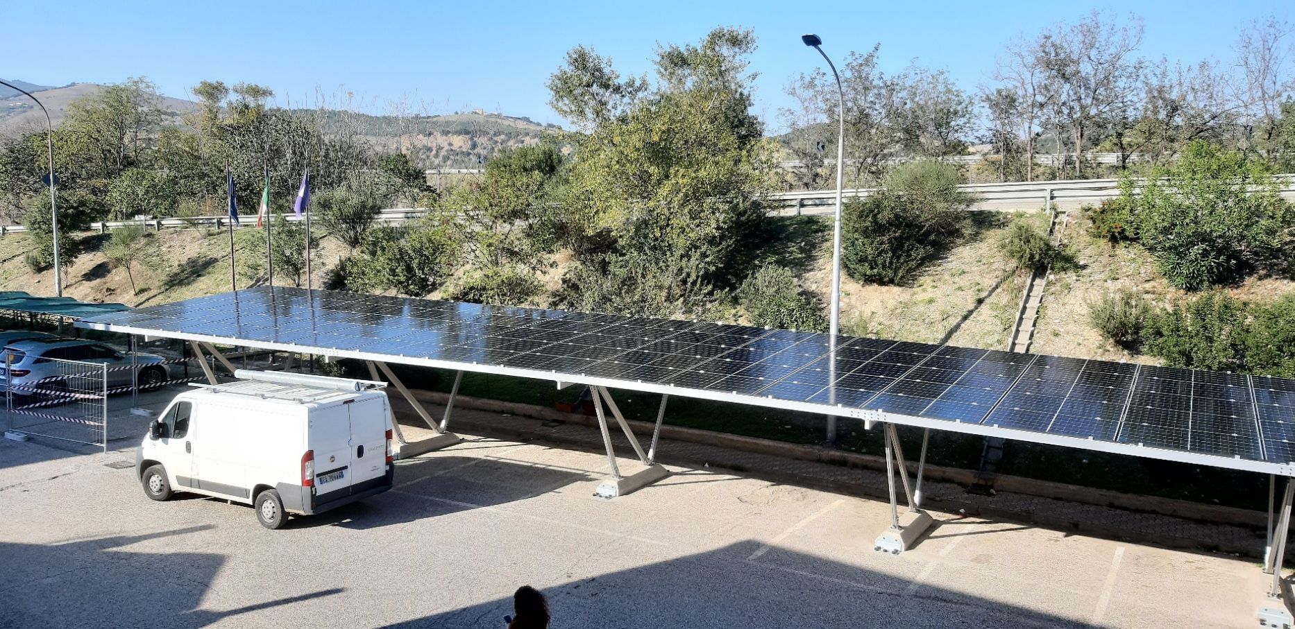 Un parcheggio con pannelli solari che creano ombra, un furgone bianco parcheggiato sotto e alberi sullo sfondo.