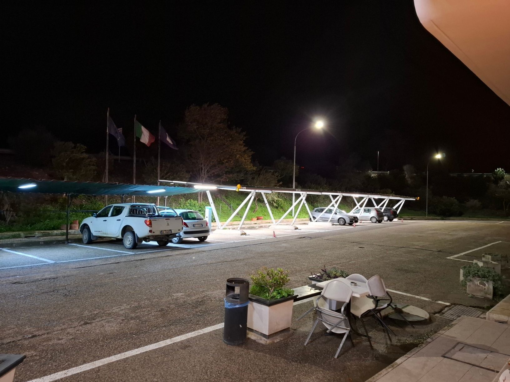 Parcheggio di notte con auto sotto una tettoia, illuminato dai lampioni. Bandiere sullo sfondo.