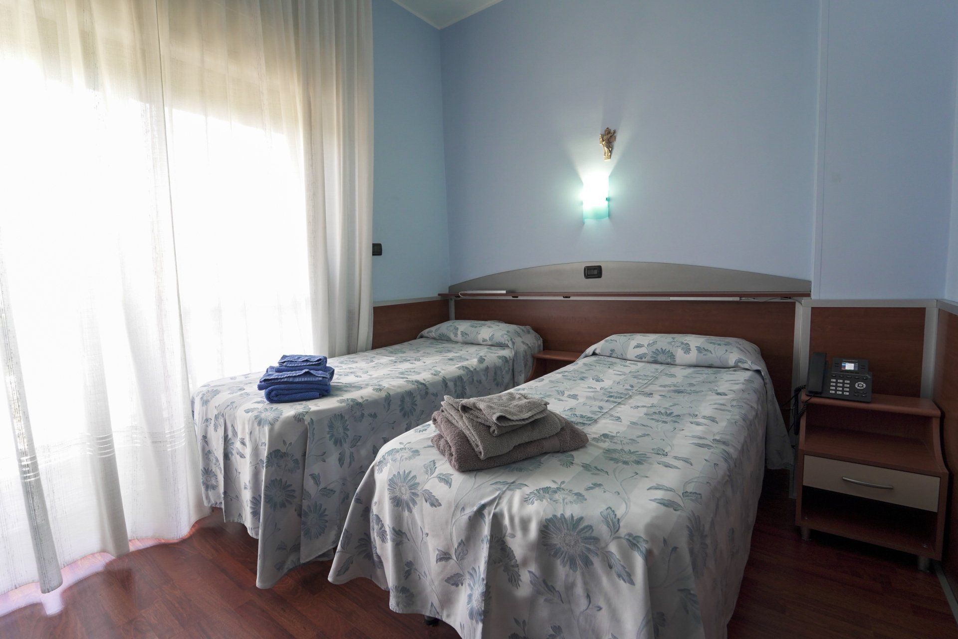 Camera d'albergo con due letti singoli. Due letti con copriletti a motivi floreali, pareti blu, una finestra con tende e un comodino.