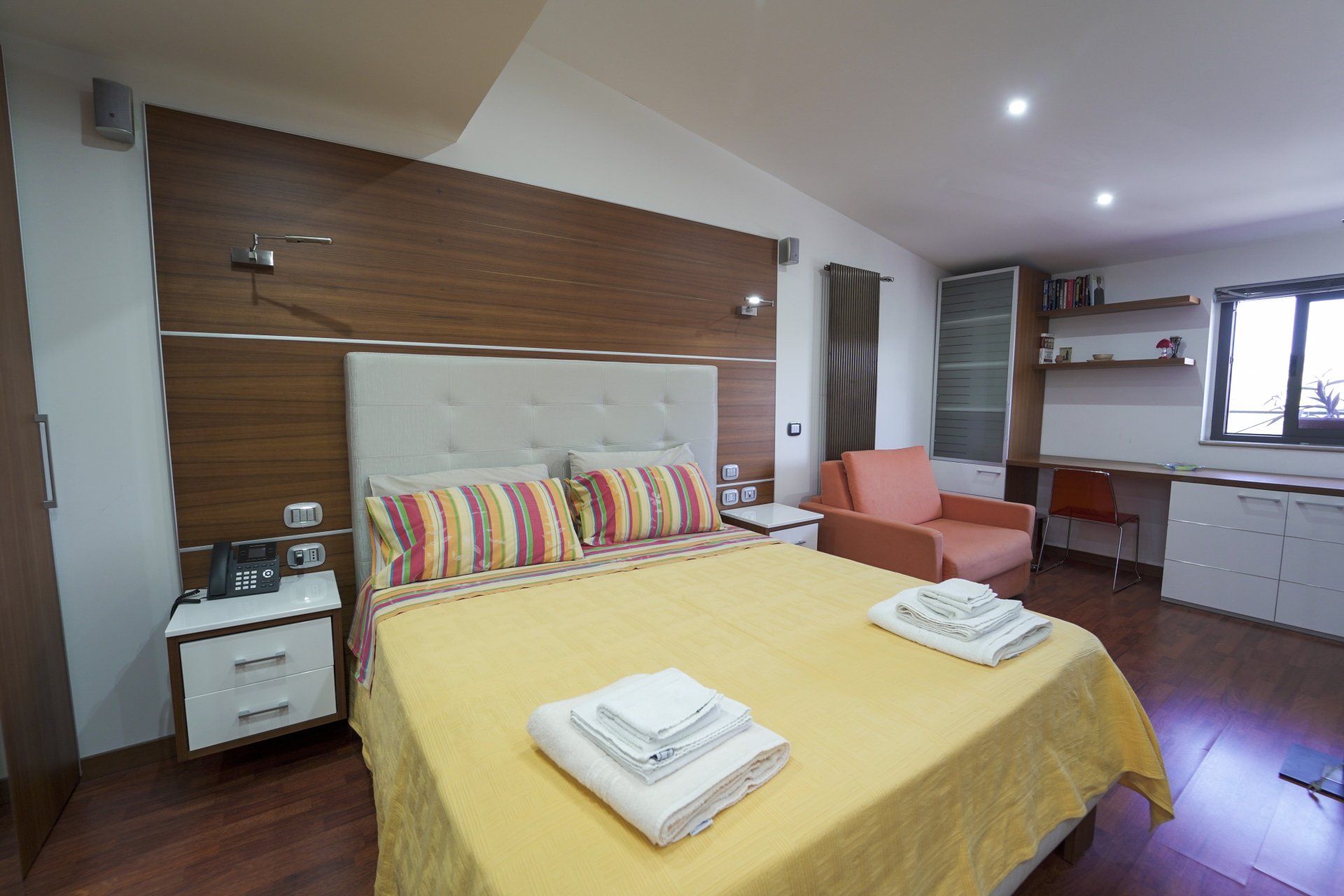 Camera da letto con letto, comodino, scrivania e poltrona. Biancheria da letto gialla e testiera in legno.