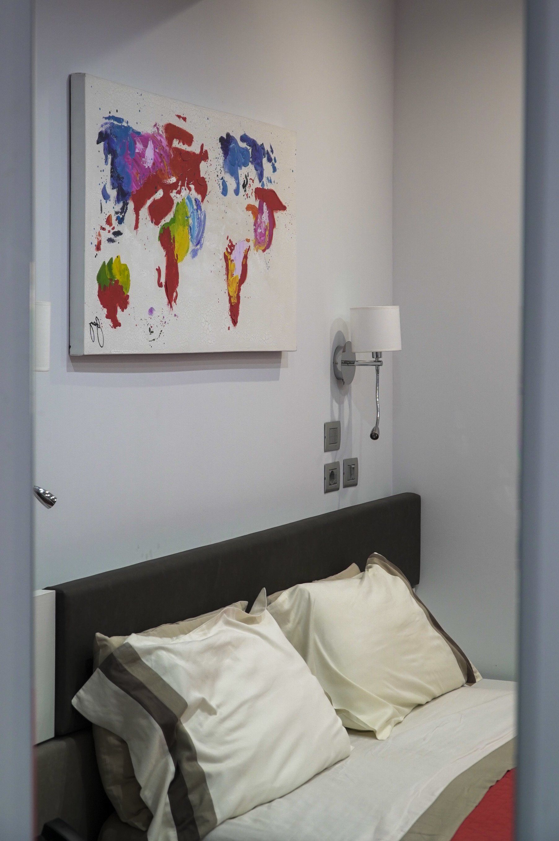 Camera da letto con letto, cuscini e un colorato dipinto raffigurante una mappa del mondo sulla parete. Accanto al letto c'è un'applique.