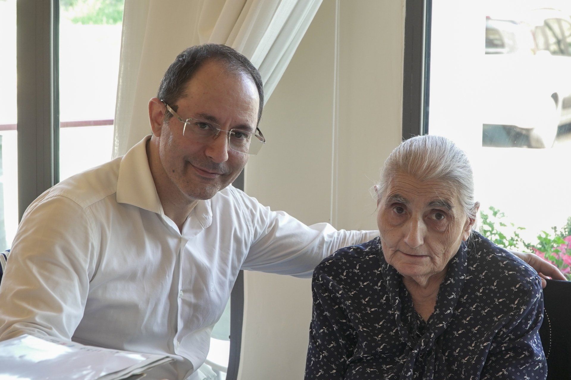 Un uomo con gli occhiali sorride, con il braccio sulla spalla di una persona anziana seduta, in casa, vicino a una finestra.