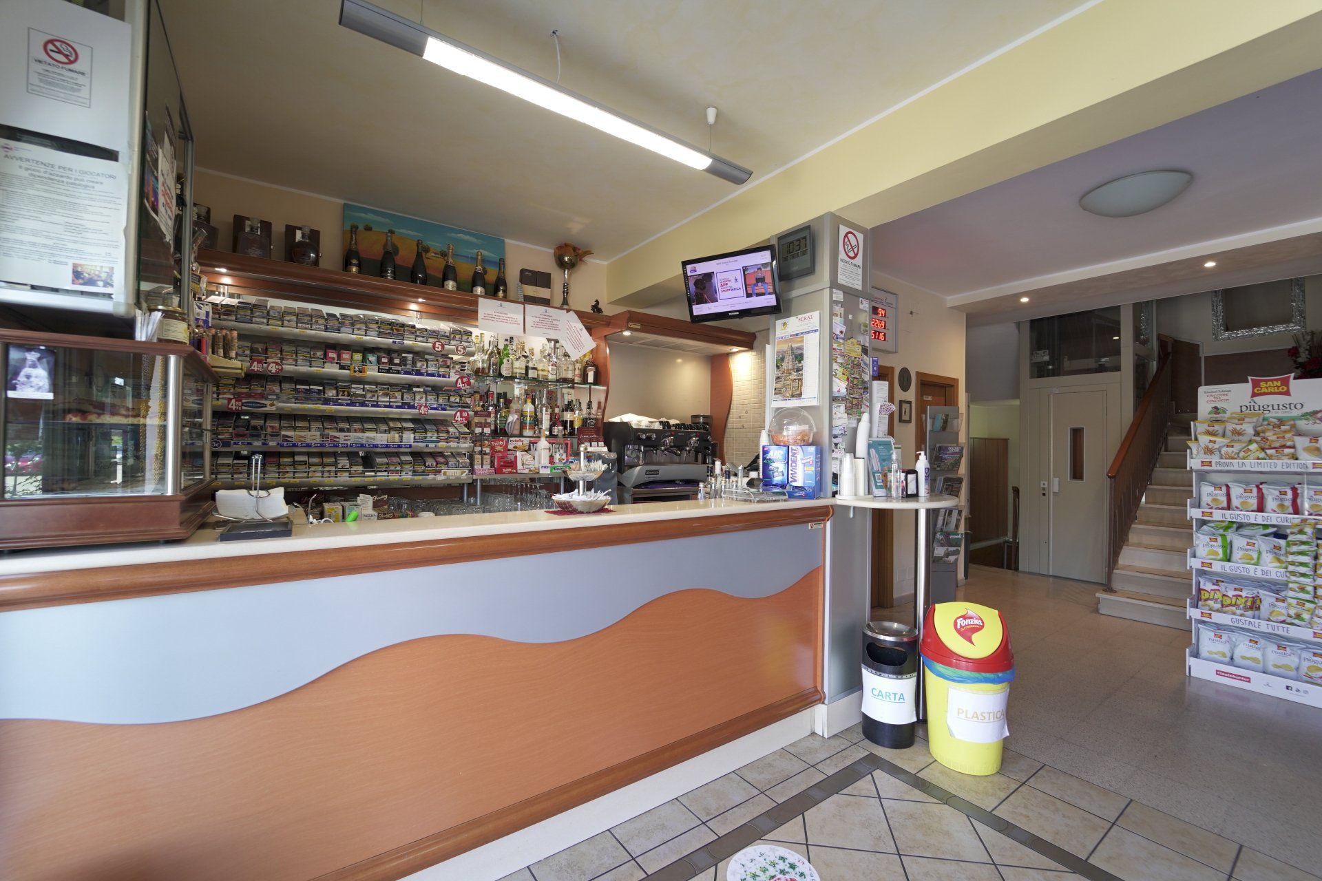 L'interno di una tabaccheria con bancone, espositori per i prodotti, TV e scale che conducono a un altro piano.
