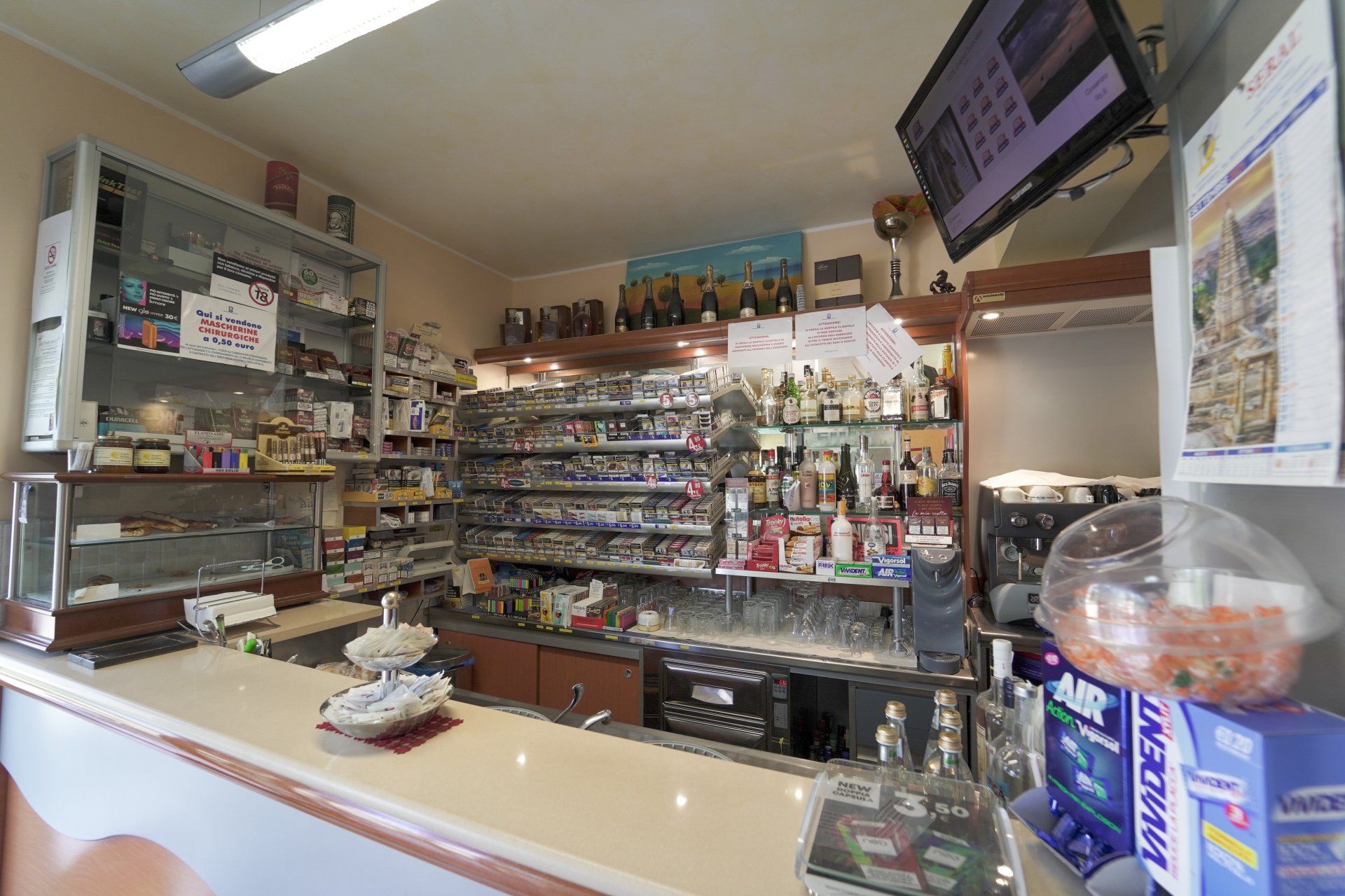 Interno di un piccolo bar con bancone e scaffali pieni di bevande e snack. È visibile uno schermo TV.