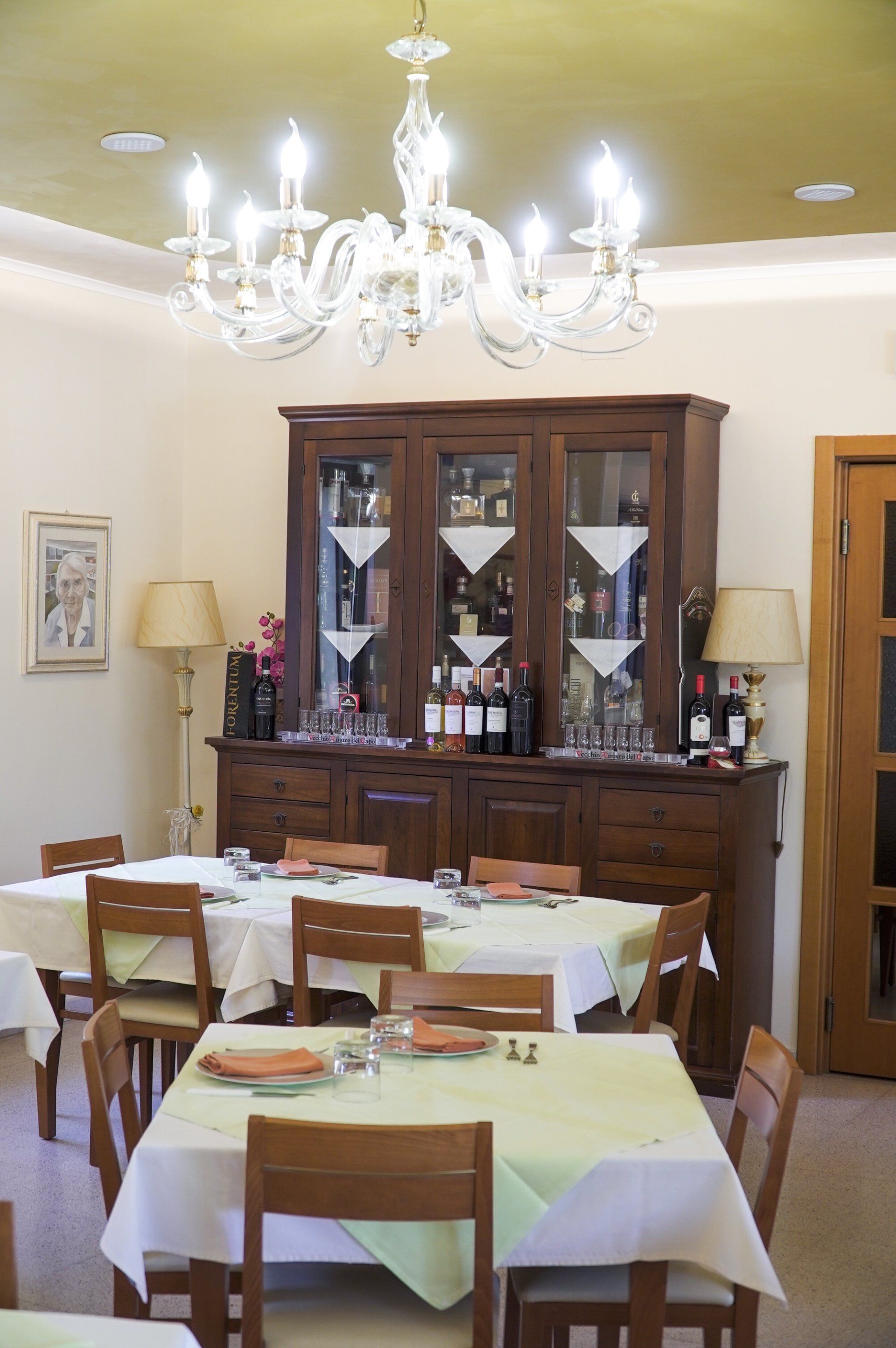 Sala da pranzo del ristorante con tavoli apparecchiati per i pasti, mobiletto in legno con bottiglie di vino, lampadario.