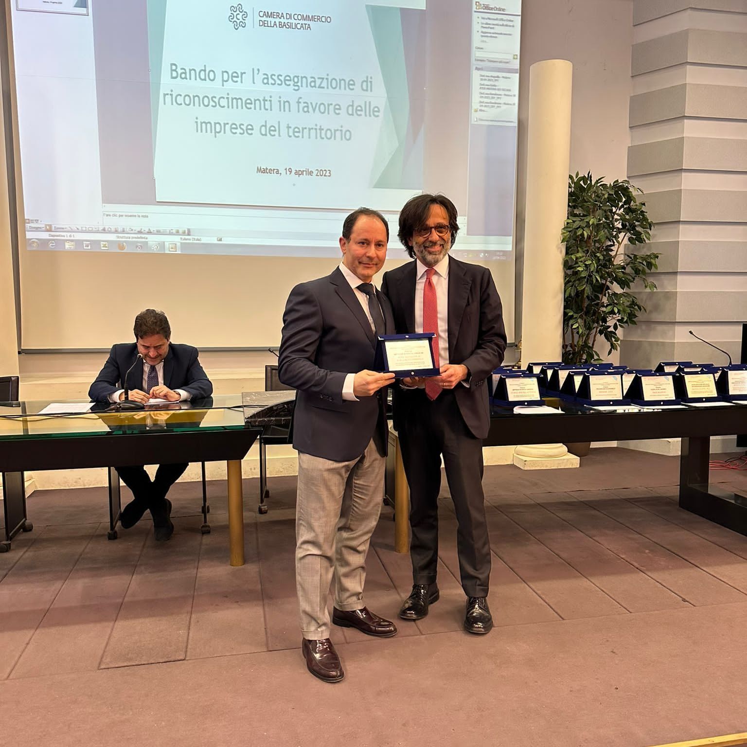 Due uomini in giacca e cravatta tengono in mano una targa durante un evento, con uno schermo di presentazione sullo sfondo.