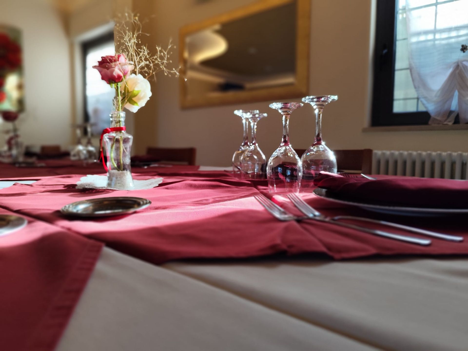 Tavolo da pranzo formale apparecchiato con tovaglia bordeaux, bicchieri e fiori in vaso.