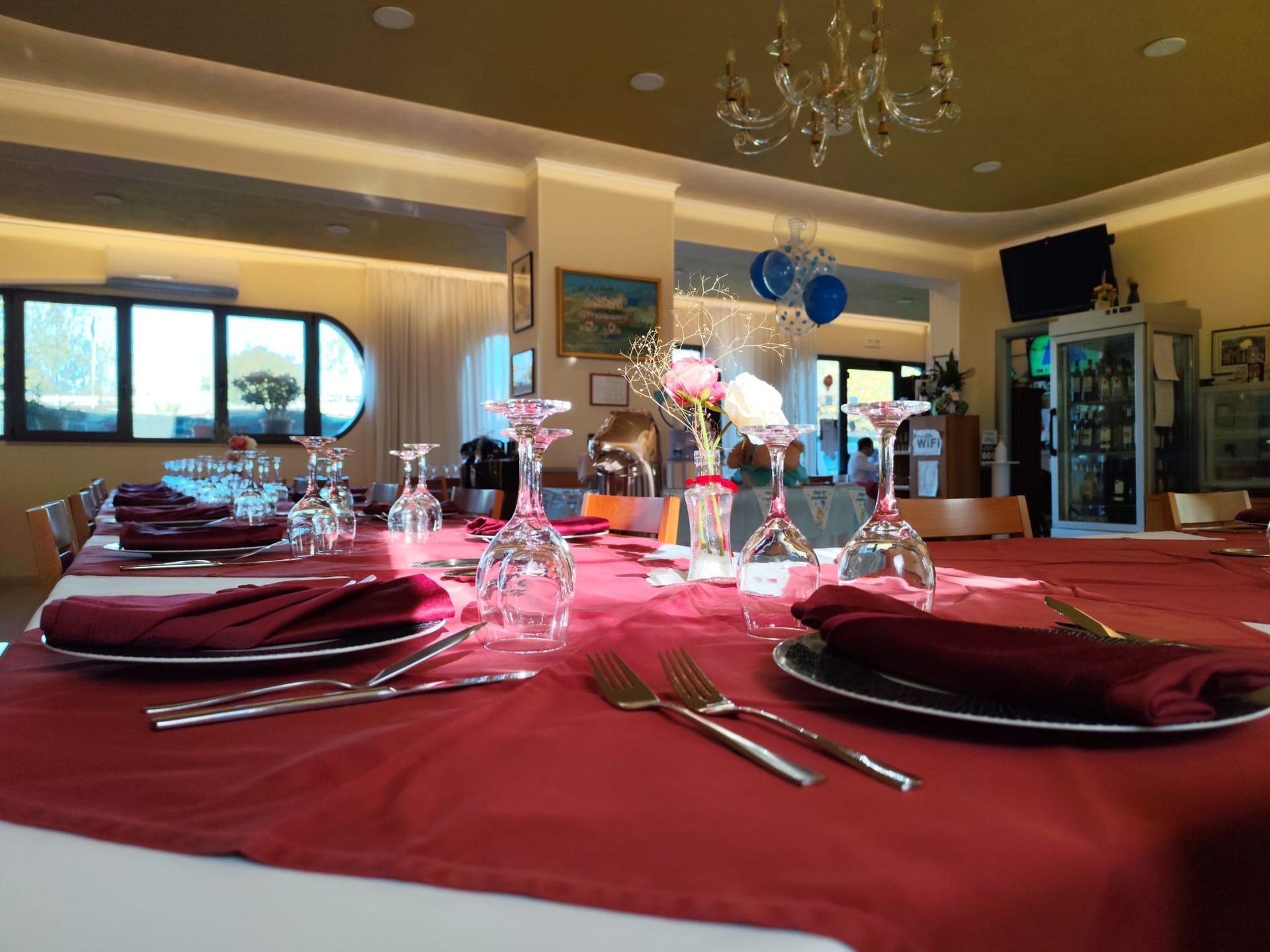 Tavolo del ristorante apparecchiato con tovaglia rossa, bicchieri da vino, posate e centrotavola; illuminazione intensa.