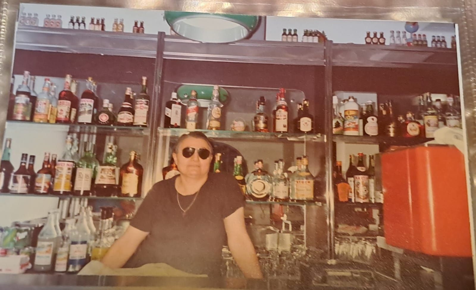Un barista con gli occhiali da sole è in piedi dietro un bancone pieno di bottiglie di liquore.