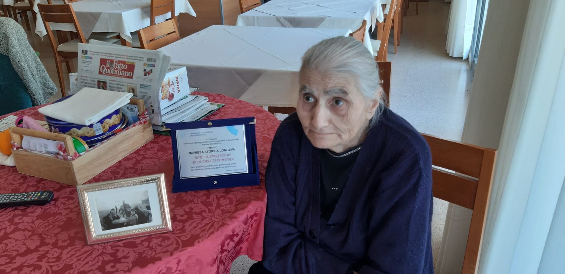 Una persona anziana è seduta a un tavolo con una foto incorniciata, una targa e una scatola di legno. Una tovaglia rossa in una stanza.