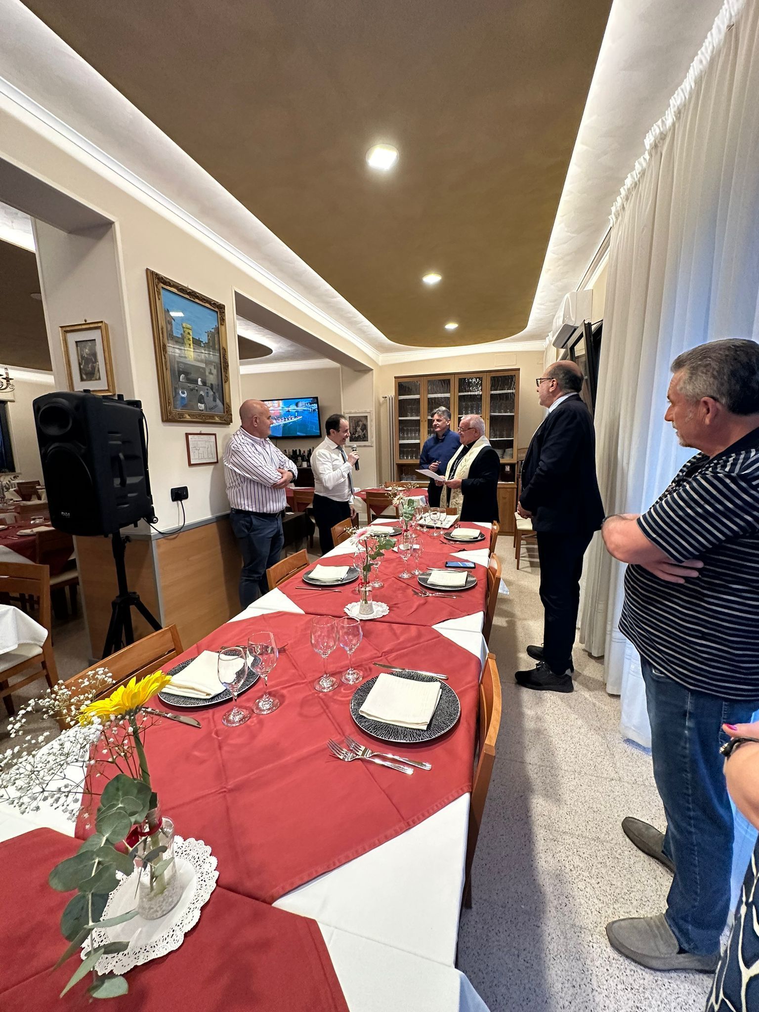 Persone riunite a un tavolo apparecchiato per un pasto in un ristorante. Un uomo parla, altri ascoltano. Tovaglia rossa.