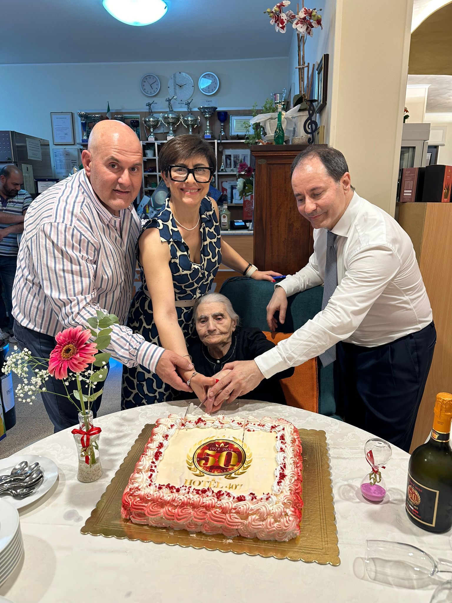 Quattro persone tagliano una torta di compleanno decorata con una persona seduta su una sedia. Allestimento per festa, fiori.