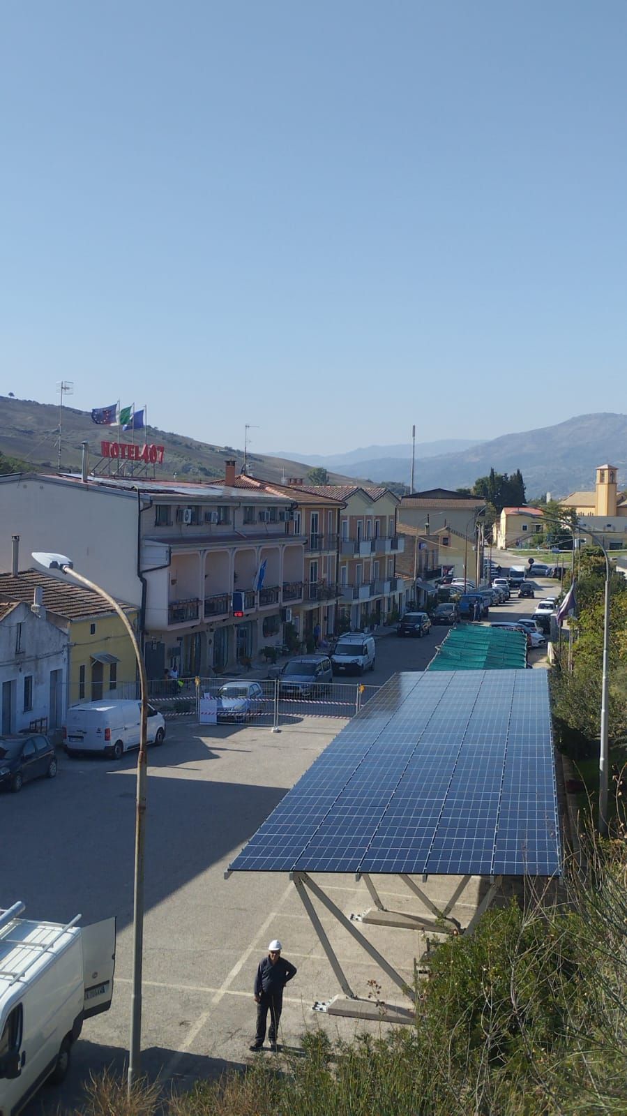 Scena di strada con tettoia di pannelli solari; auto parcheggiate lungo la strada. Edifici su entrambi i lati. Giornata limpida e soleggiata.