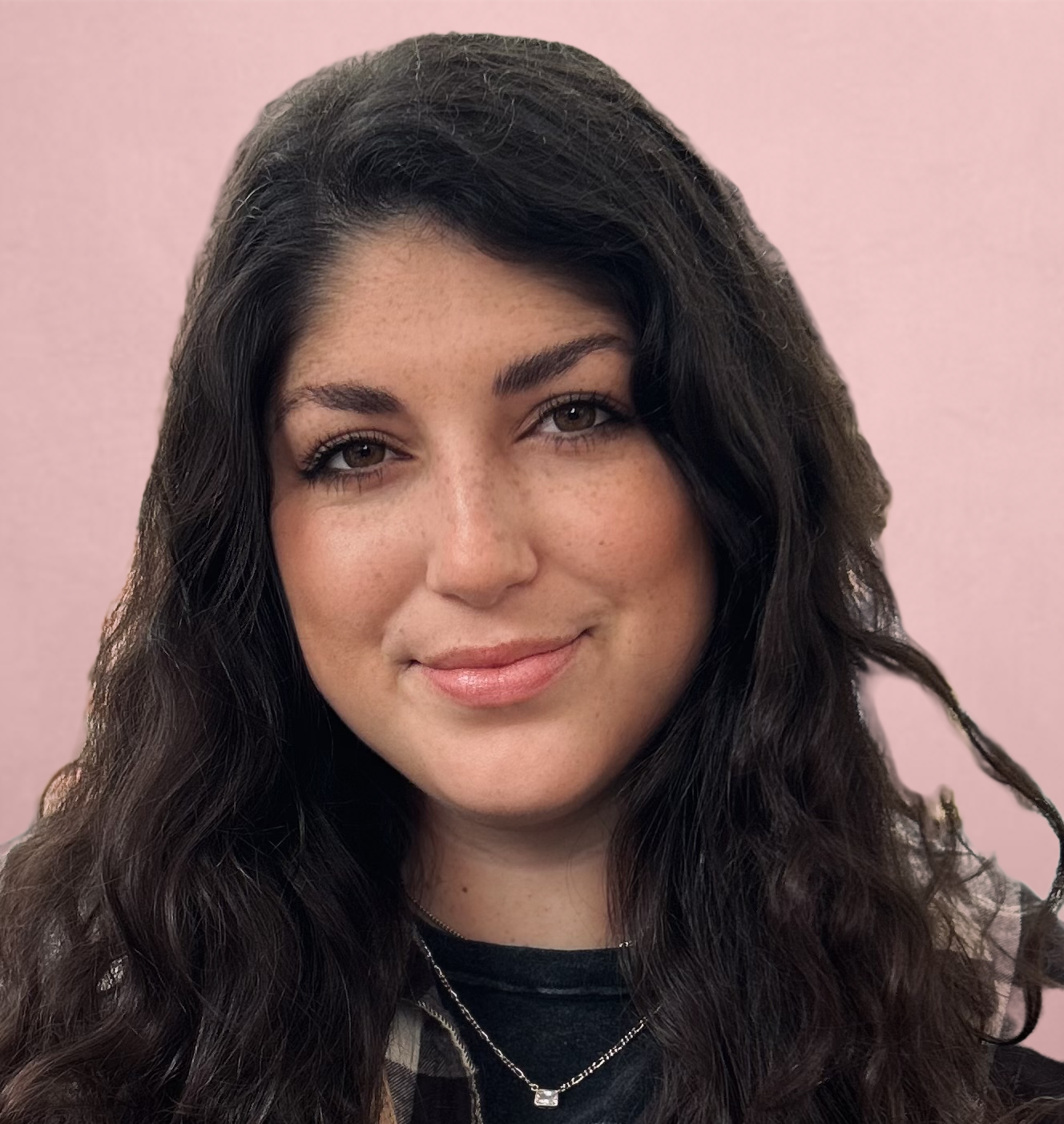 A person with long, wavy dark hair and a neutral expression stands against a light pink background.