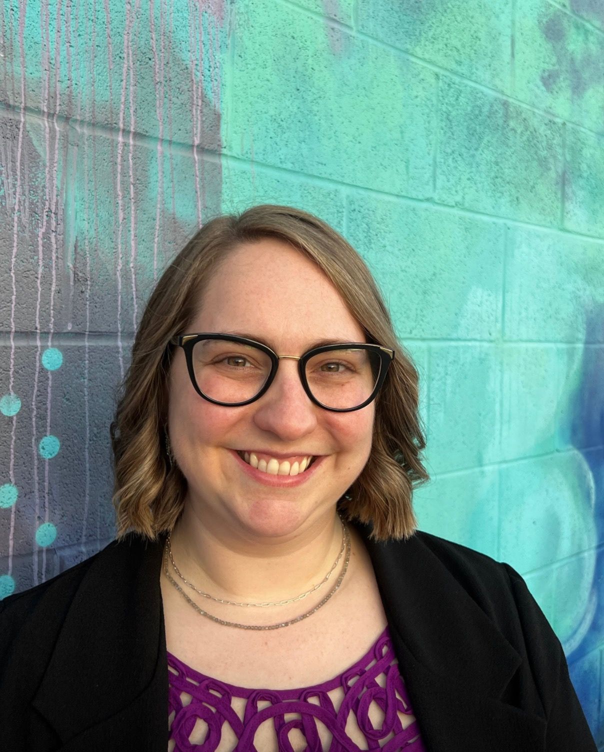 A person with shoulder-length wavy hair and glasses smiles in front of a teal and blue painted brick wall.