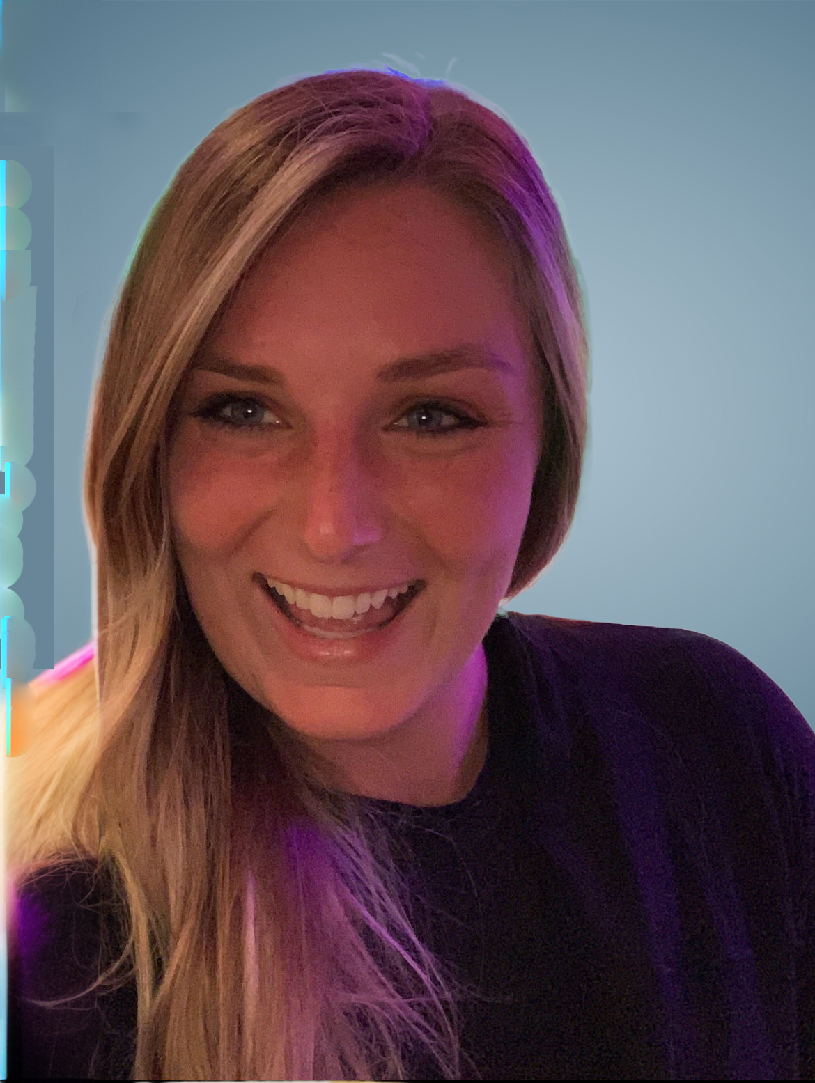 A person with blonde hair smiling brightly, wearing a dark purple top against a soft, blue-toned background.