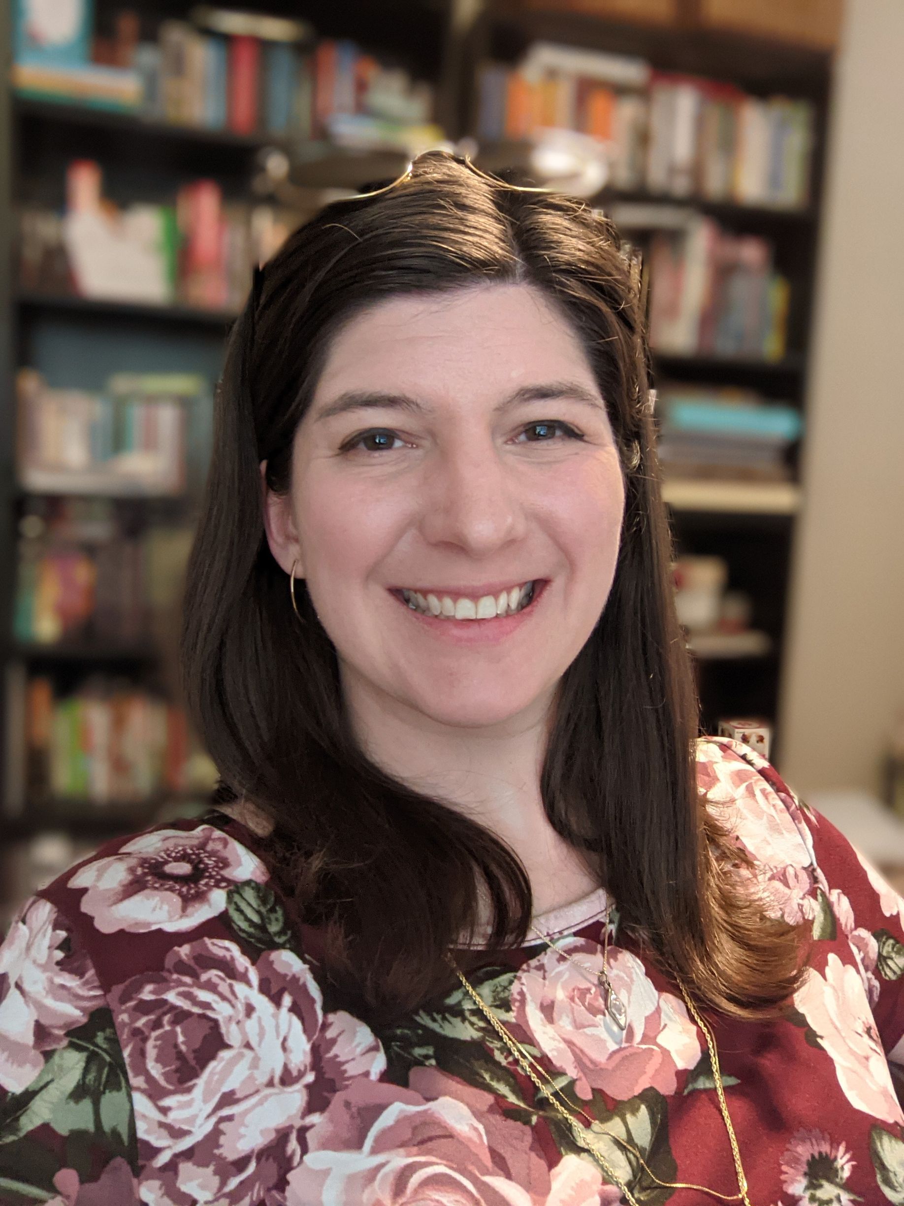 A smiling person with shoulder-length dark hair wearing a maroon floral shirt, positioned in front of a bookshelf.