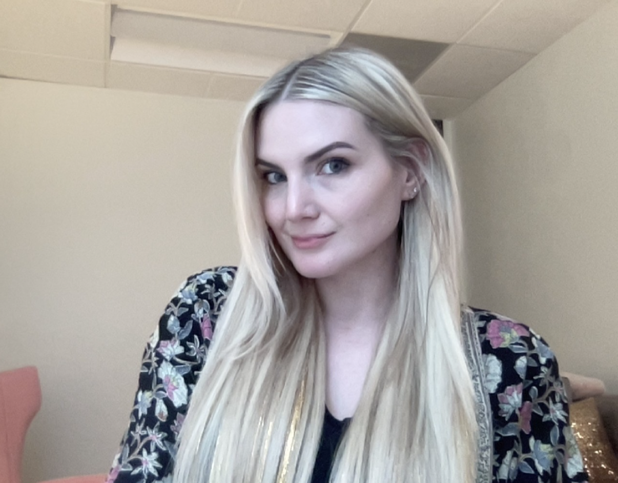 A person with long, straight blonde hair smiles gently while wearing a black floral-patterned garment in an indoor setting.