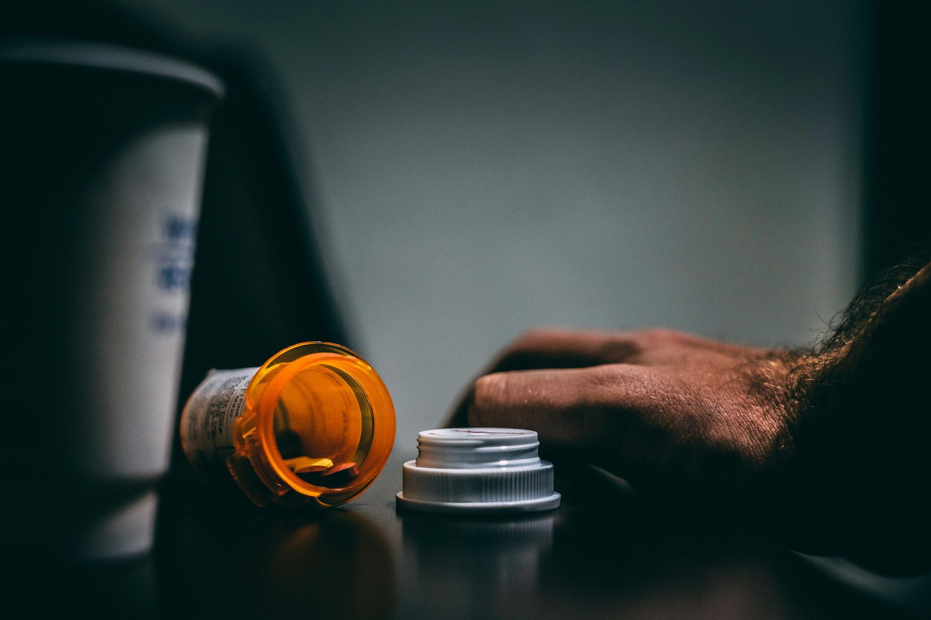 Open pill bottle, hand, and coffee cup on a dark surface.