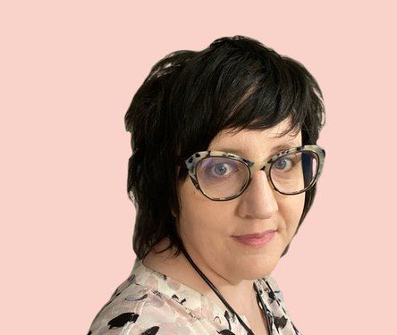 A person with short dark hair wearing patterned glasses and a light-colored printed top against a pale pink background.