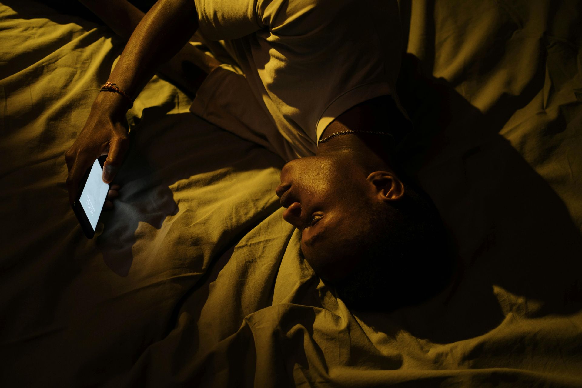 Person lying on bed, looking at lit phone screen in dim light.