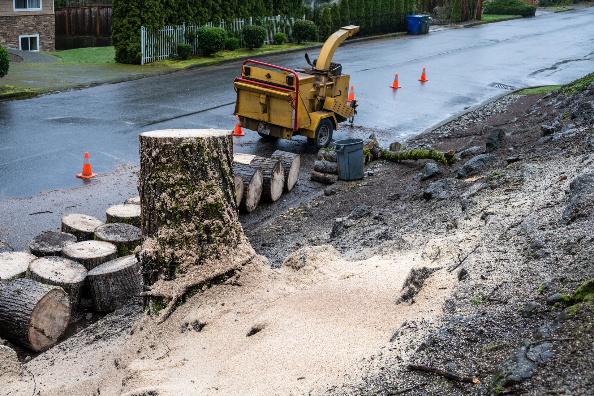 A residential street with a stump, rounds and branches alongside an industrial shredder.