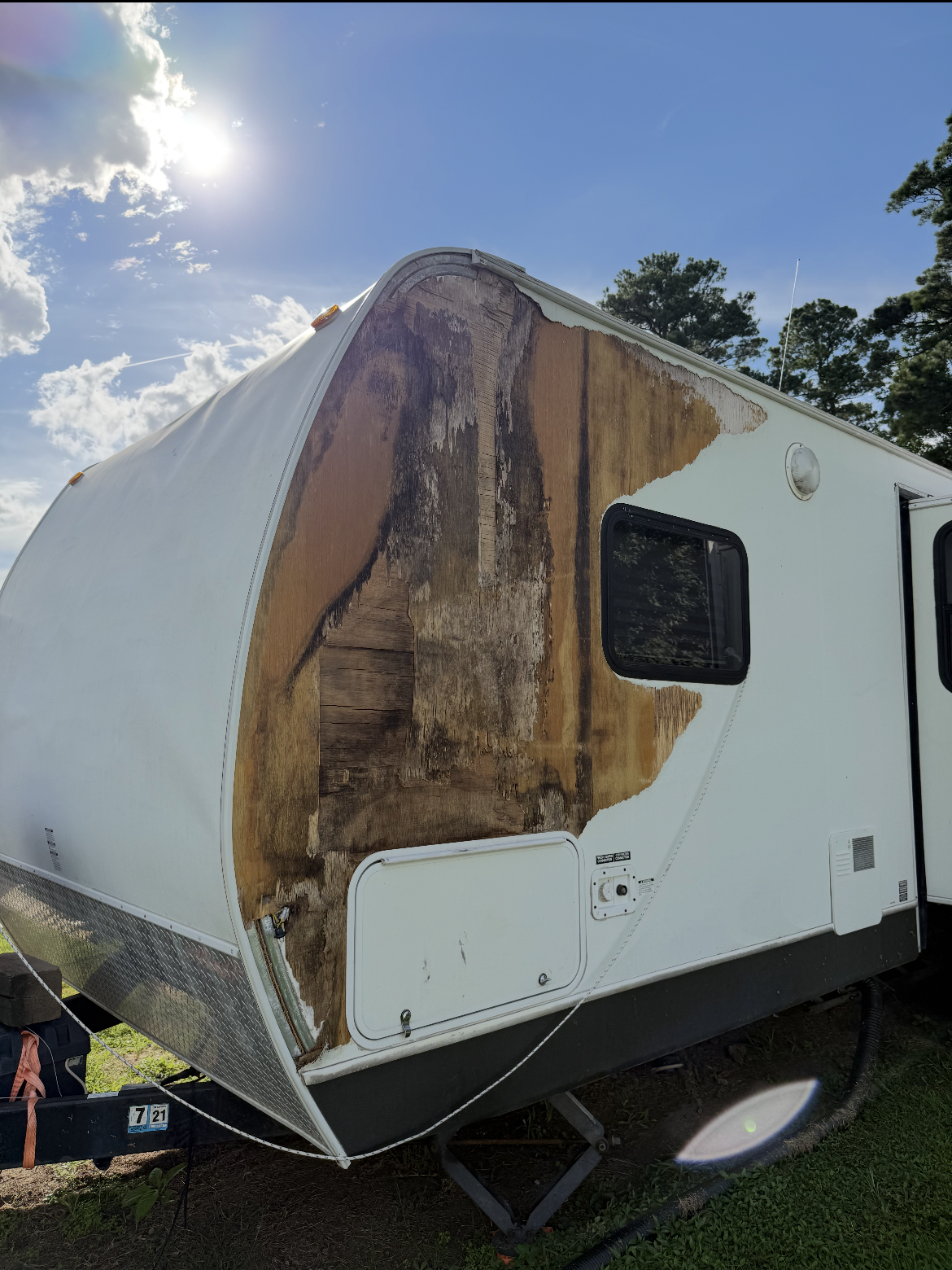 Damaged white RV trailer with a large peeled, water-stained side panel under a bright sky