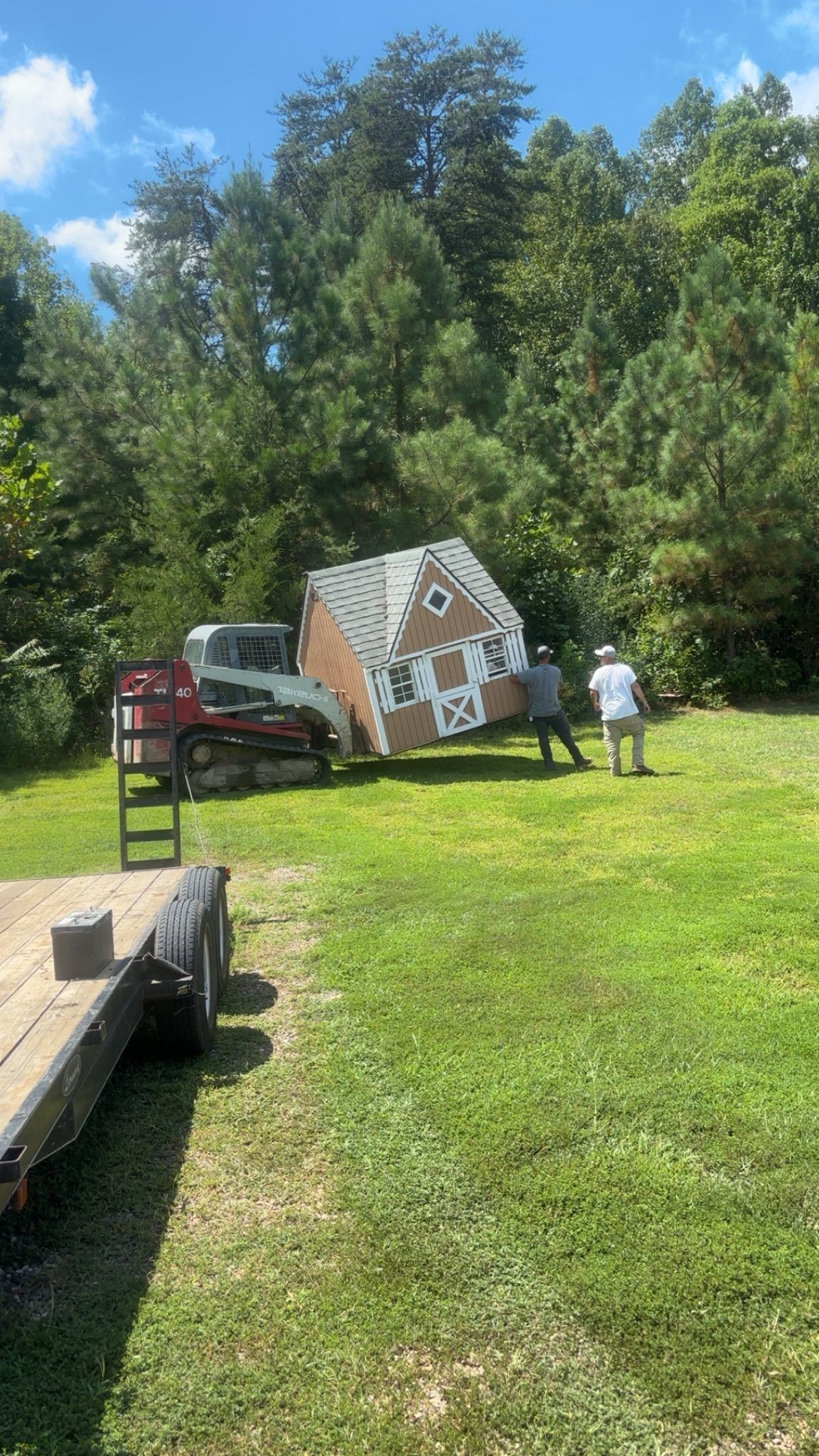 A skid steer loader moves a small, gingerbread-style house across a grassy field as two people walk beside it.